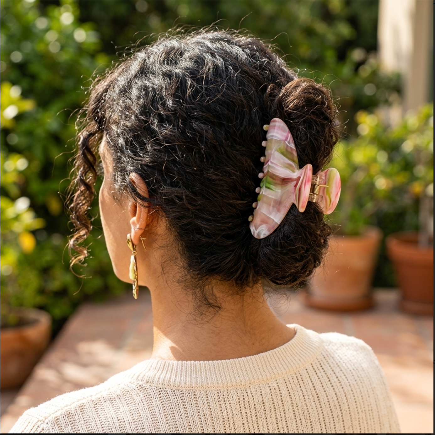 A woman shown from the back wearing a light pink claw hair clip with green and white marble-like veining, holding her dark curly hair in a bun. She is wearing a cream knit sweater and gold hoop earrings, set against a blurred outdoor garden background with potted plants.