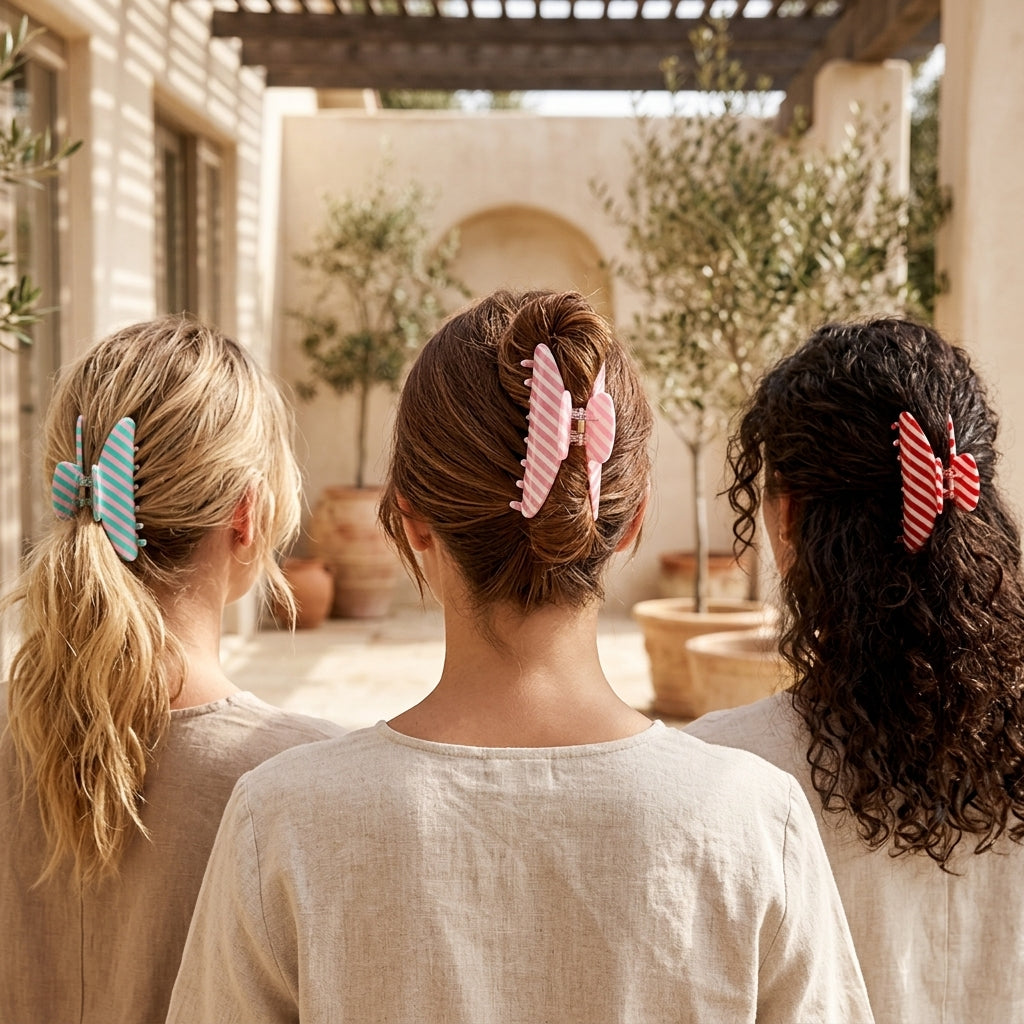 Three women seen from behind in a sunlit courtyard, each wearing a striped hair claw clip in light blue, pink, and red. The clips are styled in their hair and are the product being showcased.