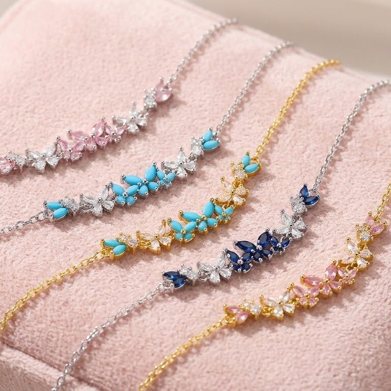 Four delicate floral chain bracelets with colorful gemstone accents are arranged on a soft pink jewelry box. The bracelets feature silver, gold, and rose gold chains with blue, pink, and dark blue flower clusters.