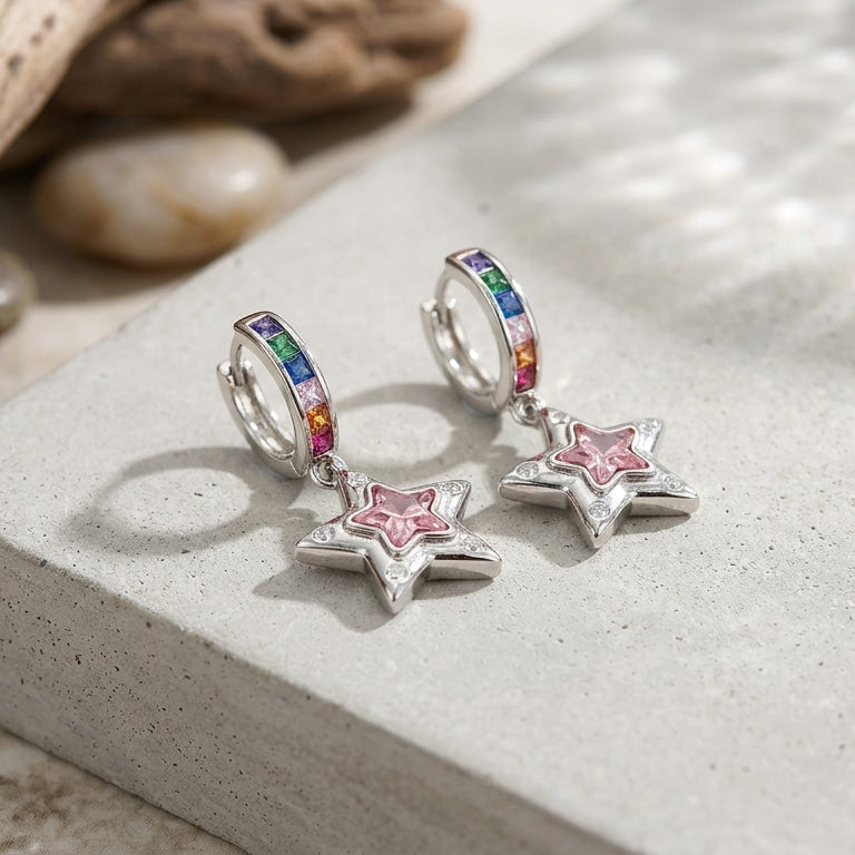 A pair of silver huggie hoop earrings with a rainbow-colored top band and dangling pink star charms, displayed on a light gray stone slab with driftwood and pebbles.
