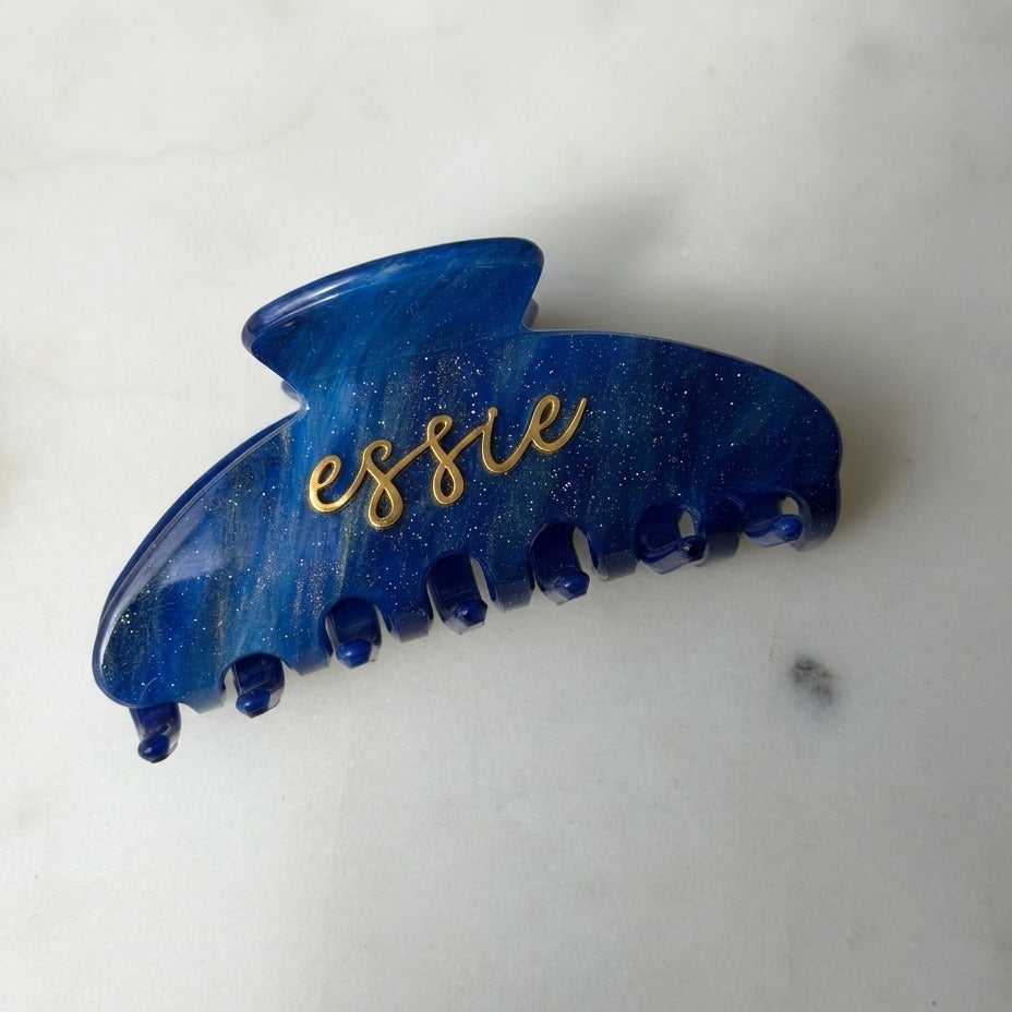 A blue marbled hair claw clip with gold glitter and the name 'essie' written in gold cursive script, resting on a light marble surface.