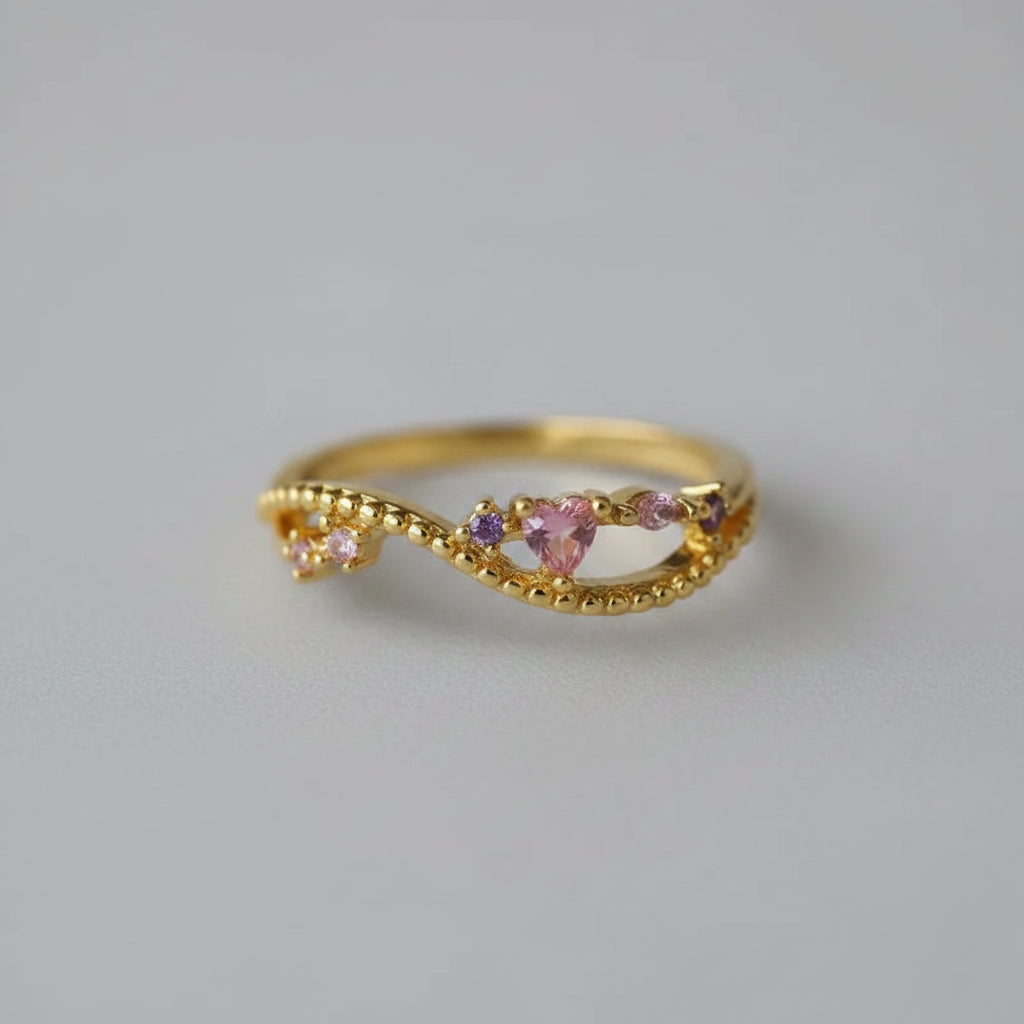 A gold-plated open ring with a twisted vine design, featuring a central heart-shaped pink gemstone and small purple accent stones, resting on a soft white fabric background.