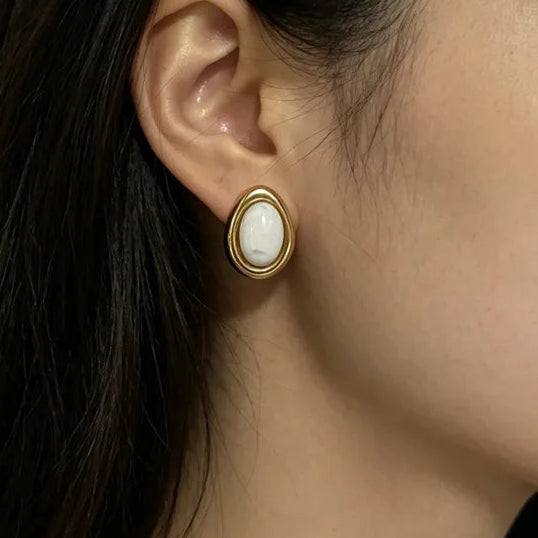 Close-up of an ear wearing a gold earring with a white stone on a neutral background