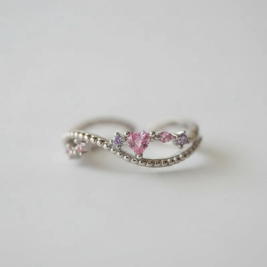 A silver-toned split ring with a heart-shaped pink gemstone and smaller pink and purple accent stones, displayed on a soft white fabric surface.