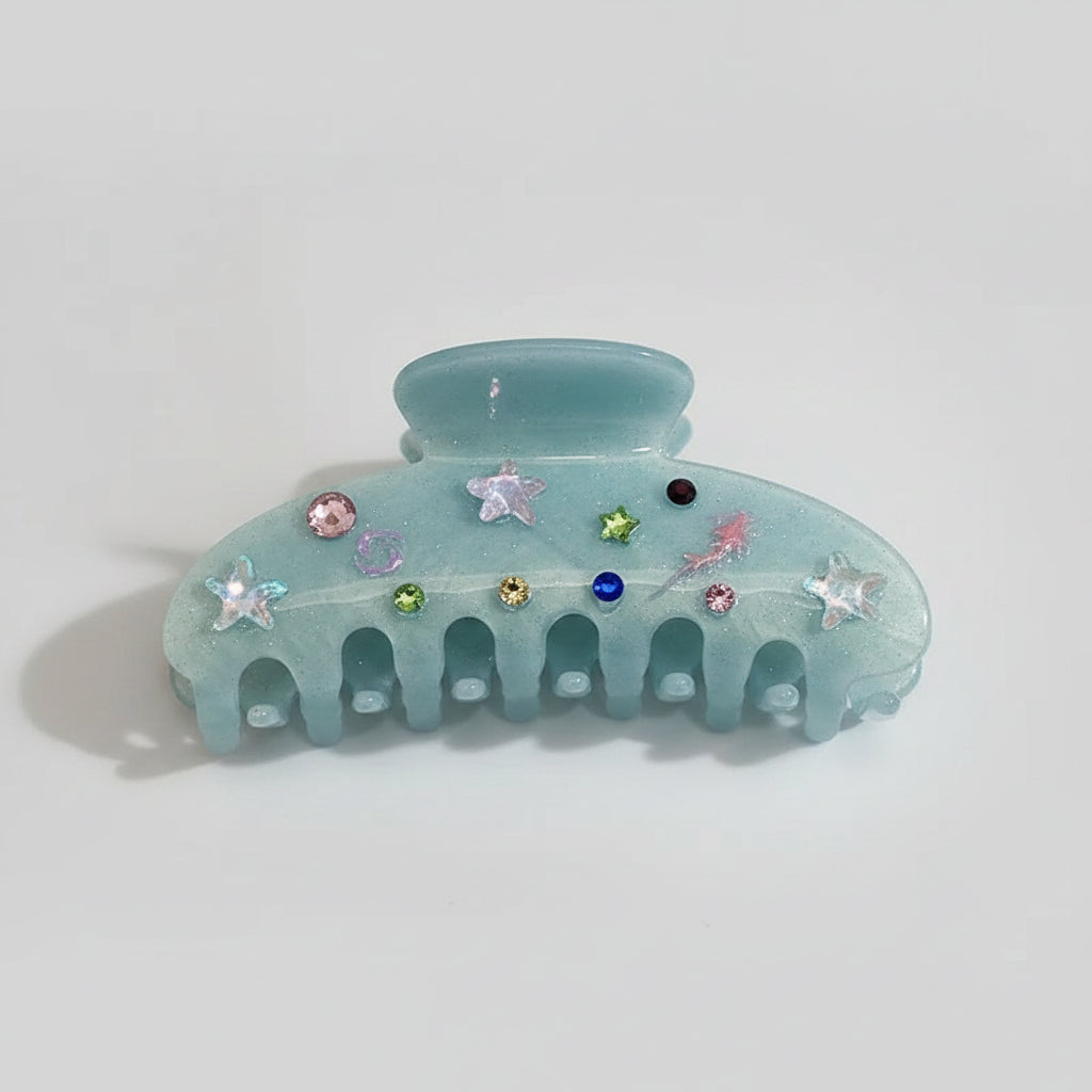 A translucent light blue-green claw hair clip decorated with small iridescent and colored rhinestones, resting on a glossy white surface with soft reflections.