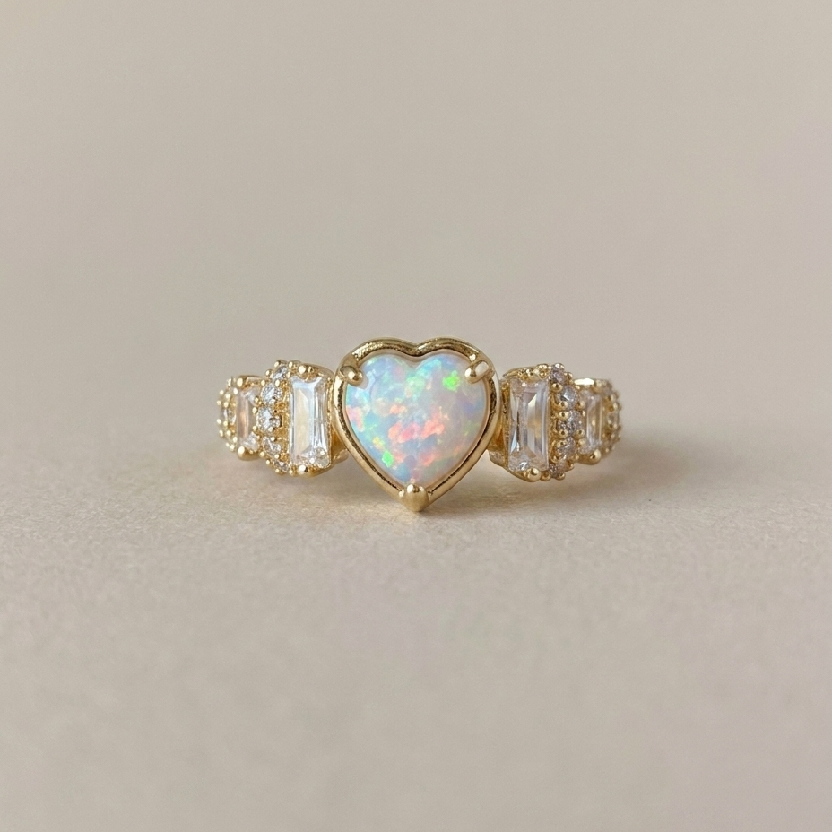 A gold ring featuring a central heart-shaped opal with iridescent white, blue, and pink flashes, flanked by baguette and round clear stones on a warm beige background.